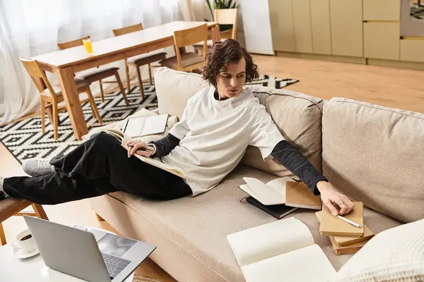 Young man comfortably sits on the couch, surrounded by books and notes, focused on studying. — Stock Photo