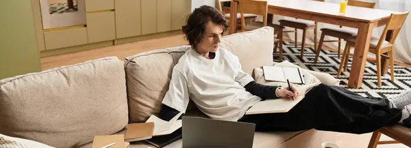 A young man sits comfortably on a couch, focused on his studies with multiple notebooks around him. — Foto stock