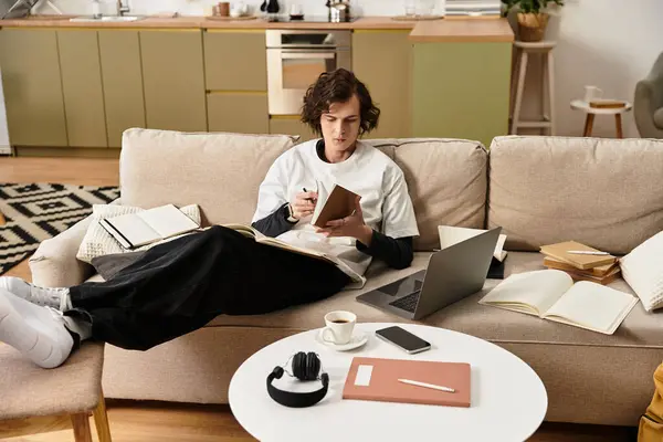 A young man relaxes on his couch, writing in a notebook with a laptop nearby and coffee in hand. — Stock Photo