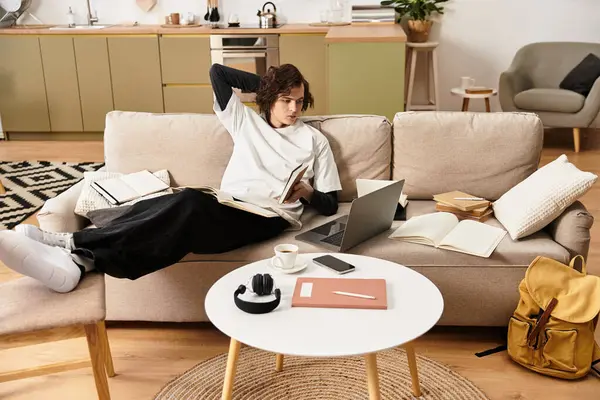 Young man in casual wear relaxes at home, reading and using his laptop among books. — Stockfoto