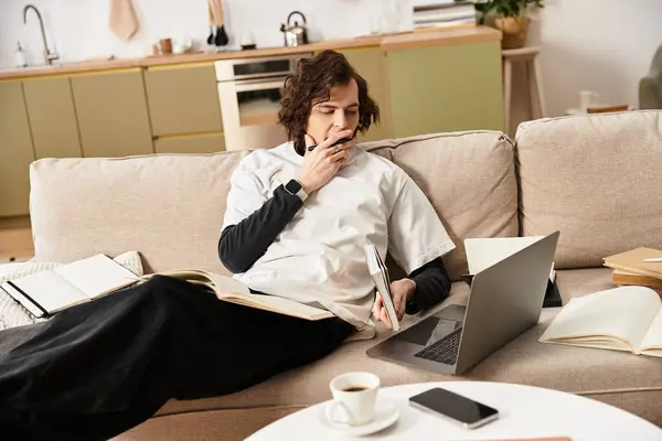 Giovane uomo si rilassa a casa, perso nel pensiero mentre multitasking con un computer portatile e libri. — Foto stock