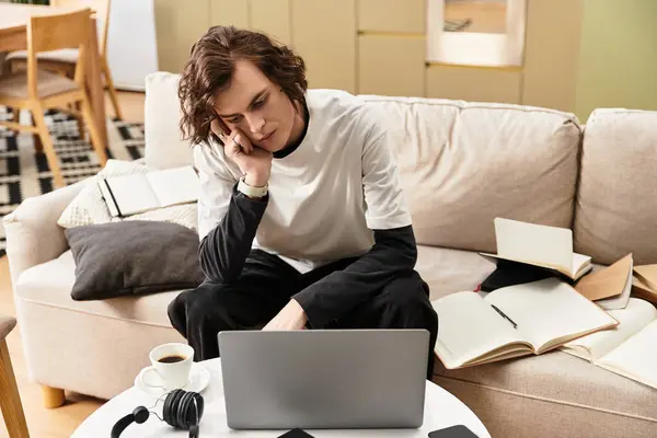 A young man sits relaxed on a sofa, focused on his laptop, surrounded by cozy comforts and books. — Fotografia de Stock