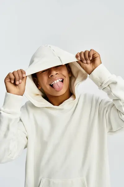 Un niño alegre muestra juguetonamente su lengua mientras usa una sudadera con capucha blanca con estilo, exudando felicidad. - foto de stock