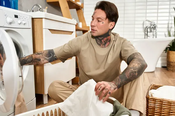 Ein gutaussehender tätowierter junger Mann sitzt gemütlich auf dem Fußboden und erledigt zu Hause Wäschearbeiten. — Stockfoto