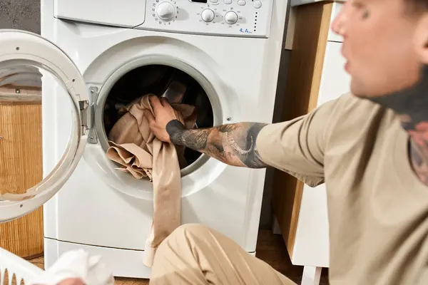 A young man with tattoos places clothes in the washing machine while relaxing at home. — Stock Photo
