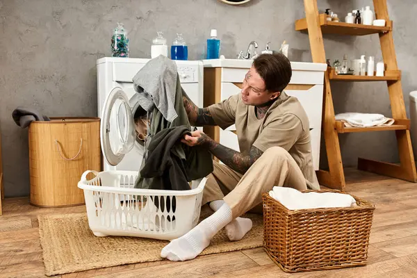 Ein tätowierter Mann sortiert Kleidung aus seinem Korb in eine Waschmaschine, während er sich zu Hause entspannt. — Stockfoto