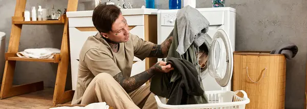 Ein junger tätowierter Mann konzentriert sich darauf, Wäsche in eine warme, einladende Waschmaschine zu laden. — Stockfoto