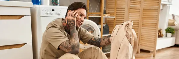 A young tattooed man sits thoughtfully in his warm home, surrounded by laundry and comfort. — Stock Photo