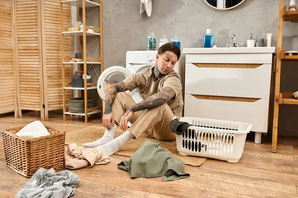 Ein junger tätowierter Mann sitzt gemütlich auf dem Boden und sortiert Wäsche in einer gemütlichen häuslichen Umgebung. — Stockfoto