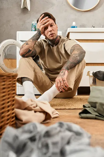 Ein tätowierter junger Mann entspannt auf dem Boden, umgeben von Wäsche in einem gemütlichen Haus. — Stockfoto