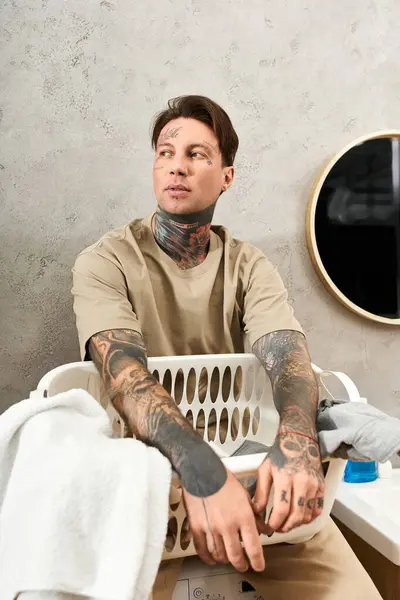 A young tattooed man creates a cozy vibe as he relaxes with a laundry basket. — Stock Photo
