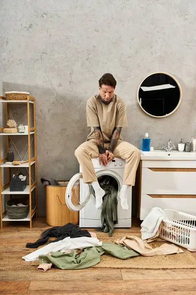 Ein gutaussehender Mann mit Tätowierungen entspannt zu Hause an der Waschmaschine, umgeben von Wäsche. — Stockfoto
