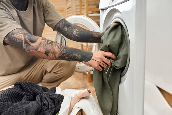 Ein junger Mann mit Tätowierungen genießt einen entspannten Moment beim Falten von Wäsche in einem warmen, einladenden Raum. — Stockfoto
