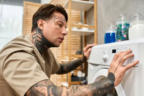 Ein junger tätowierter Mann, der sich auf das Waschen von Kleidung konzentriert, umgeben von einem gemütlichen, modernen Wohnraum. — Stockfoto