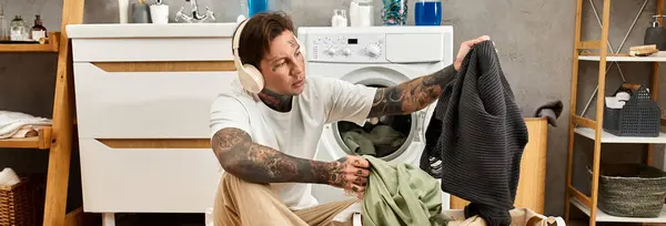 Ein tätowierter junger Mann entspannt zu Hause, wäscht Wäsche und genießt Musik mit Kopfhörern. — Stockfoto