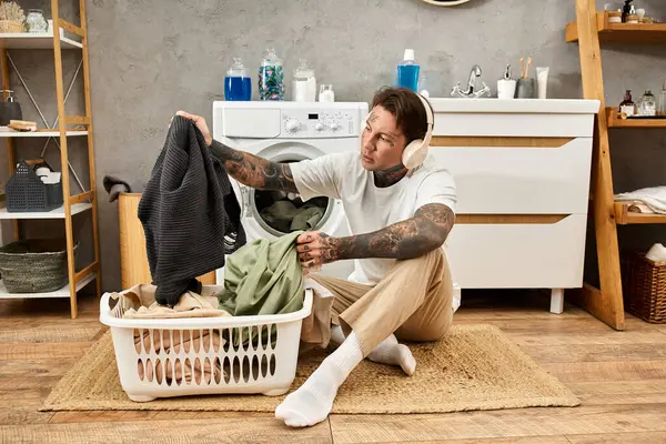 Young man with tattoos enjoys a comfy day at home while sorting laundry and listening to music. — Stock Photo