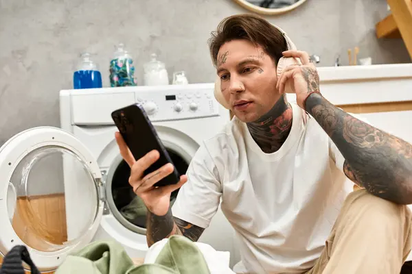 Tätowierter junger Mann hört Musik über Kopfhörer und entspannt sich zu Hause neben Waschmaschinen. — Stockfoto