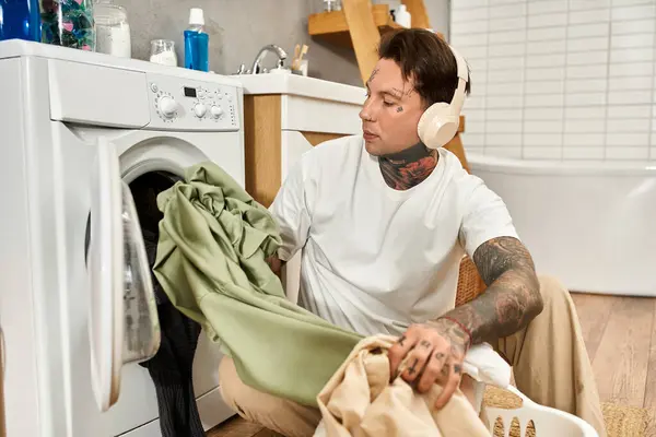 Junger Mann mit Tätowierungen geht zu Hause fröhlich Wäsche waschen und genießt einen entspannten Moment. — Stockfoto