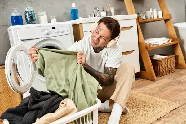 Schöner tätowierter Jugendlicher sitzt gemütlich zu Hause und sortiert Kleidung aus der Waschmaschine — Stockfoto