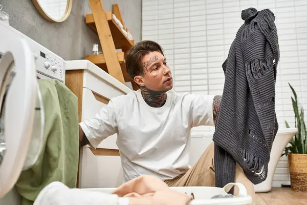 Ein gut aussehender tätowierter Mann verbringt Zeit zu Hause, sortiert Wäsche und genießt dabei eine gemütliche, entspannte Atmosphäre. — Stockfoto
