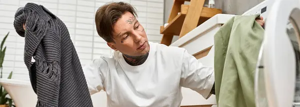 Ein junger Mann mit Tätowierungen konzentriert sich auf Wäschearbeiten und balanciert Kleidungsstücke in einer gemütlichen Umgebung. — Stockfoto