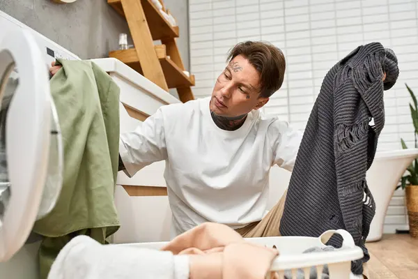 Young man with tattoos sorts laundry at home, surrounded by a serene and inviting space. — Stock Photo