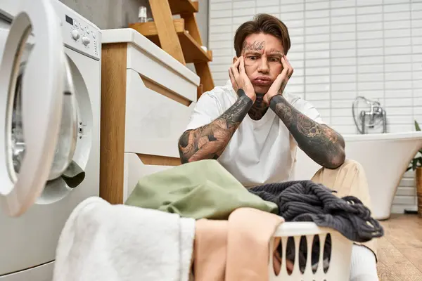 Ein tätowierter junger Mann sitzt nachdenklich neben einem Wäschekorb in seinem gemütlichen Zuhause. — Stockfoto