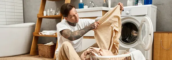 In einem gemütlichen Zuhause sortiert ein junger tätowierter Mann Wäsche, während er einen entspannten Moment genießt. — Stockfoto