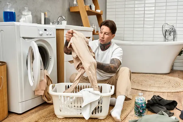 Ein junger tätowierter Mann faltet sorgfältig Wäsche, umgeben von einem gemütlichen Badezimmer. — Stockfoto