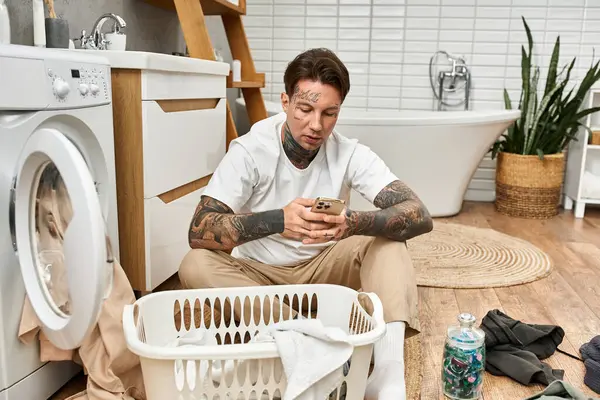 Ein junger Mann mit Tätowierungen sitzt gemütlich zu Hause, vertieft in SMS und Wäschepflichten. — Stockfoto