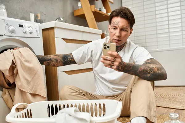 Handsome tattooed man sits comfortably folding laundry while checking his smartphone. — Stock Photo