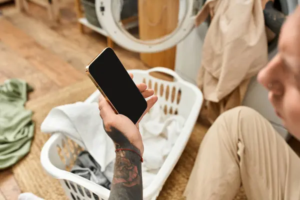 In a cozy home setting, a young tattooed man takes a break from laundry to check his smartphone. — Stock Photo