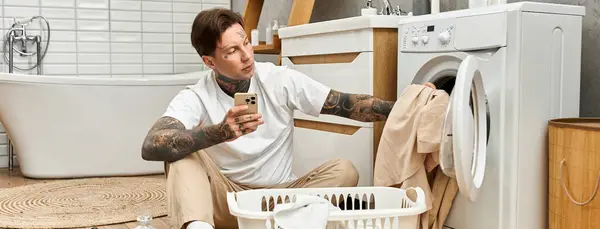 In einem gemütlichen Badezimmer entspannt sich ein tätowierter junger Mann, sortiert Wäsche und checkt sein Telefon. — Stockfoto