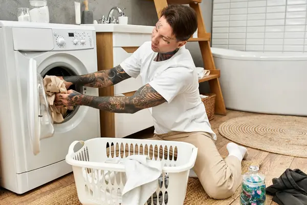 Tatuato giovane uomo carica lavanderia in una lavatrice in un ambiente rilassato. — Foto stock