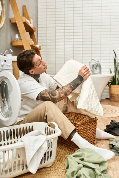 Tattooed young man sits comfortably at home while folding fresh laundry with a relaxed expression. — Stock Photo