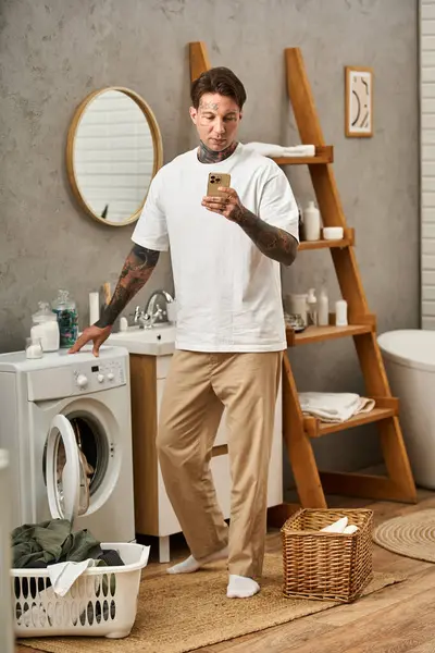 Junger Mann mit Tätowierungen entspannt sich in seiner Wohnung und checkt beim Wäschewaschen sein Handy. — Stockfoto