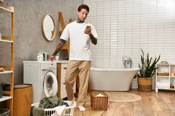 Ein stilvoller junger Mann checkt sein Handy, während er in einem hellen und luftigen Badezimmer Wäsche wäscht. — Stockfoto
