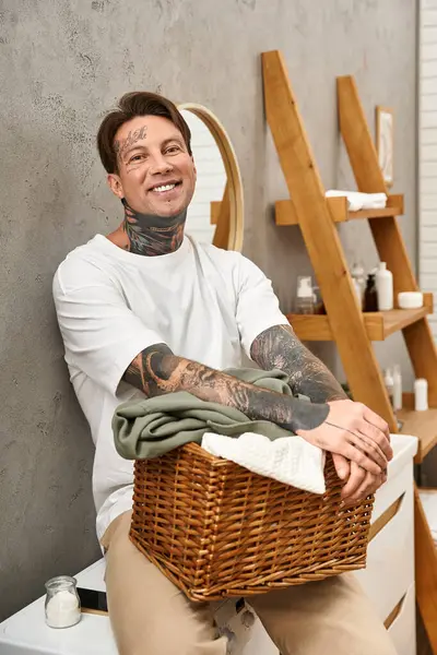 Lächelnder junger Mann mit Tätowierungen entspannt zu Hause, während er einen geflochtenen Wäschekorb in der Hand hält. — Stockfoto