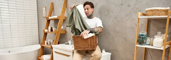 A stylish young man with tattoos sits in a relaxing bathroom, sorting laundry in a basket. — Stock Photo