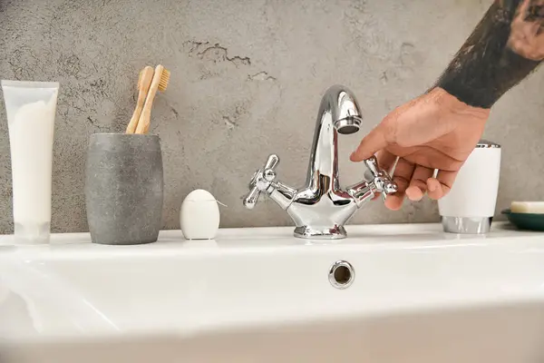 Ein junger, tätowierter Mann steht in einem gemütlichen Badezimmer und dreht mit Leichtigkeit den Wasserhahn auf. — Stockfoto