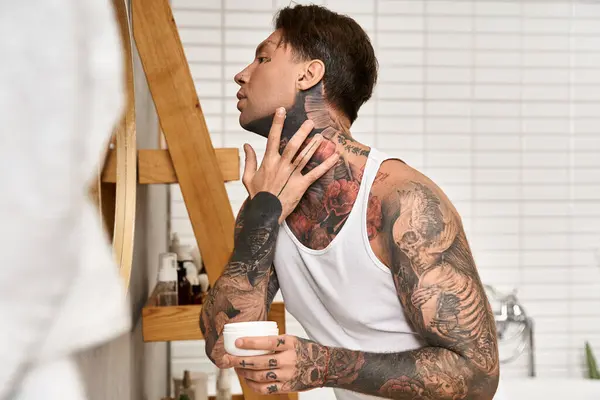 Handsome young man applies cream while admiring his tattoos in a comfortable home setting. — Stock Photo