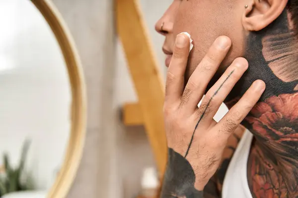 Ein junger Mann mit auffälligen Tätowierungen trägt beim Entspannen zu Hause Feuchtigkeitscreme auf sein Gesicht auf. — Stockfoto