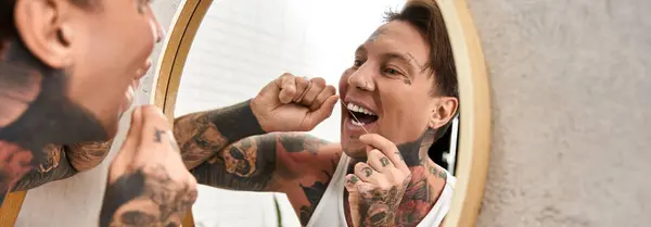 Joven guapo con tatuajes sonríe a su reflejo mientras usa hilo dental en un cómodo baño. - foto de stock