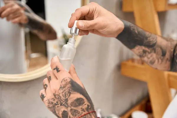 A handsome tattooed man holds a skincare bottle, enjoying a relaxed moment in his cozy home. — Stock Photo