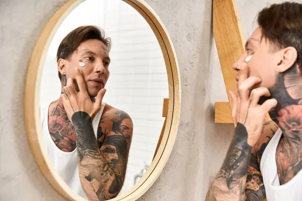 Hombre tatuado aplica crema mientras mira su reflejo en un espejo, abrazando el autocuidado en casa. - foto de stock