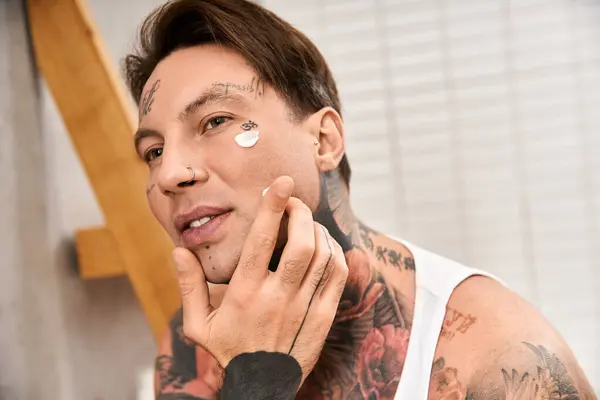 Ein tätowierter junger Mann trägt in einem gemütlichen Heim Feuchtigkeitscreme auf, umarmt die Selbstpflege. — Stockfoto