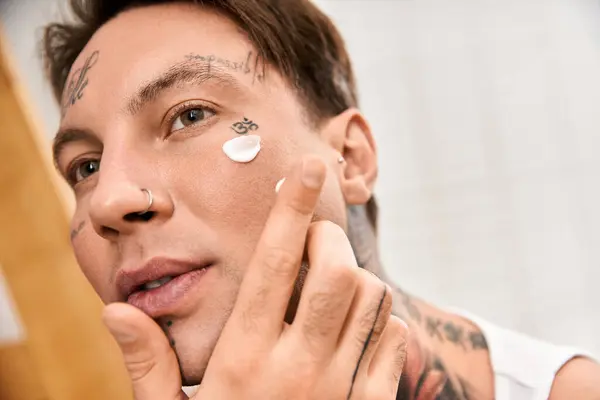 Ein junger tätowierter Mann genießt seine Hautpflege zu Hause und cremt sein Gesicht sorgfältig ein. — Stockfoto