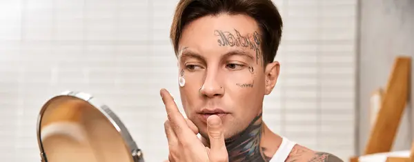 Ein junger tätowierter Mann nimmt sich Zeit für die Selbstpflege, während er im Spiegel cremt. — Stockfoto