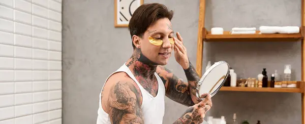 A tattooed young man applies skincare in the mirror, enjoying a moment of relaxation. — Stock Photo