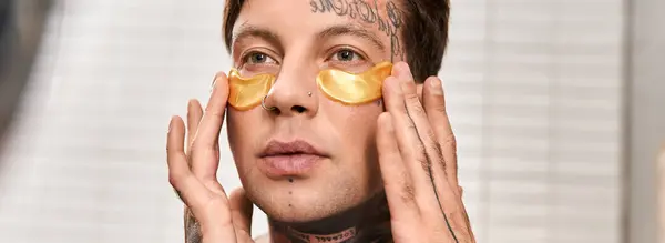 Ein junger Mann mit Tätowierungen trägt Gold unter Augenklappen auf, während er es sich in seinem gemütlichen Zuhause gemütlich macht. — Stockfoto